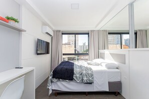 A modern bedroom with a bed, a TV, a desk, and a window with curtains. - Studio no All You Need - All0604 (Curitiba)