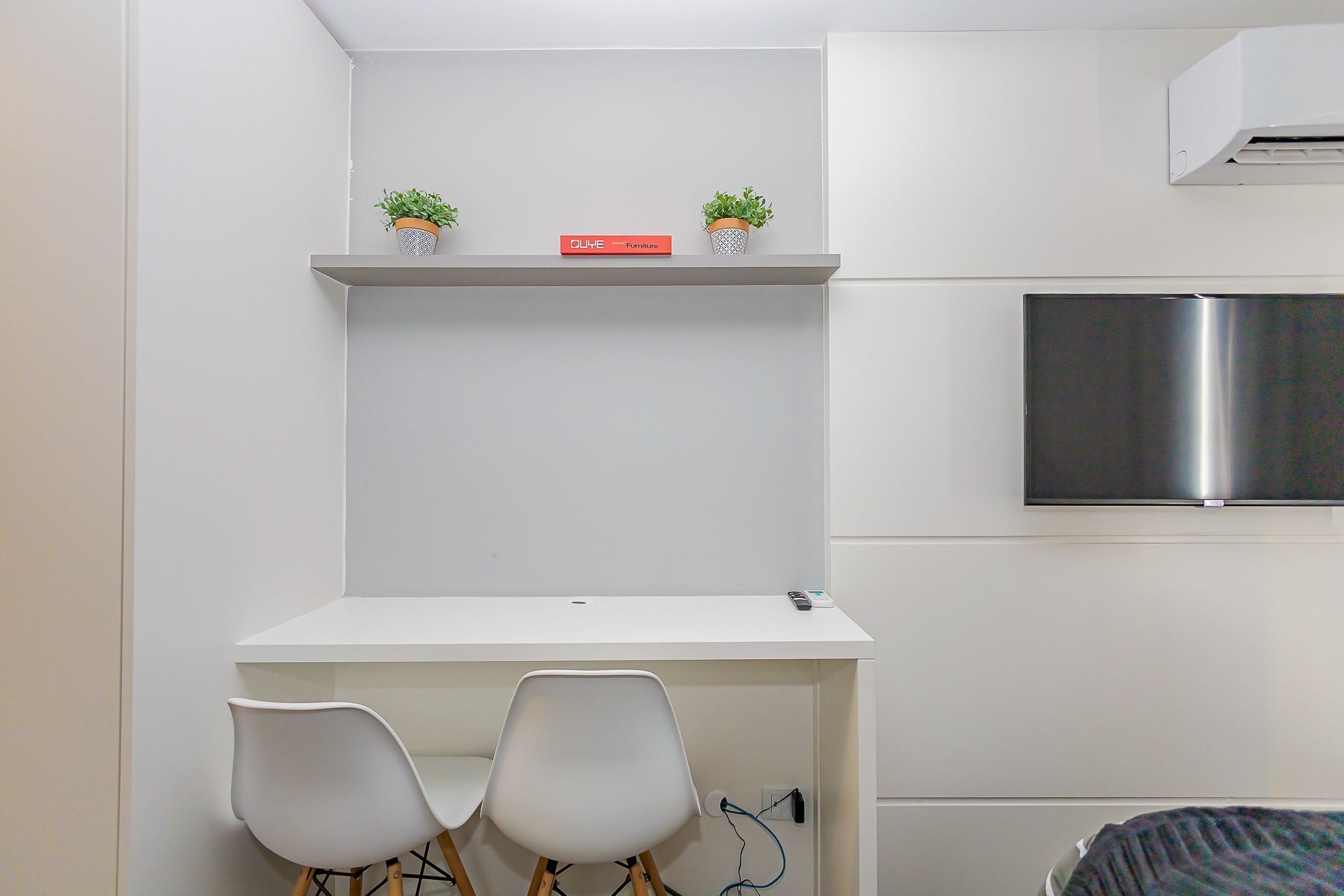 Una habitación compacta con un escritorio blanco, dos sillas blancas, un estante pequeño con dos plantas en macetas, un libro rojo titulado 