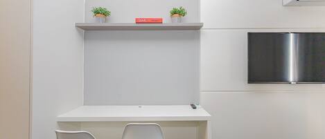 Una habitación compacta con un escritorio blanco, dos sillas blancas, un estante pequeño con dos plantas en macetas, un libro rojo titulado