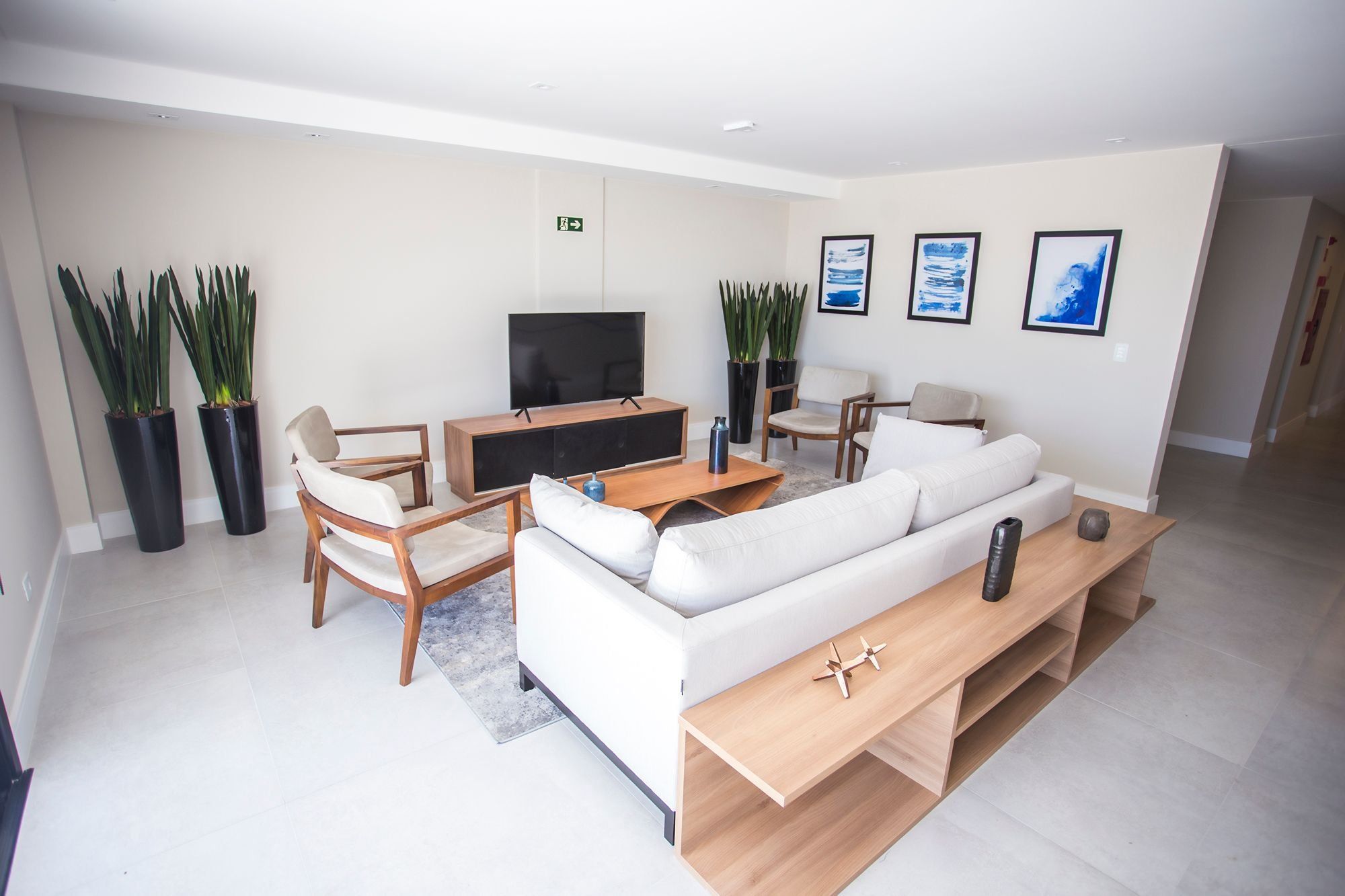 Uma sala de estar moderna com um sofá branco, mesa de centro de madeira e uma televisão de tela plana. Há duas plantas em vasos e uma pequena mesa com um controle remoto.