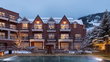 A multi-story hotel with a pool in the foreground, featuring balconies and a view of snow-covered mountains.