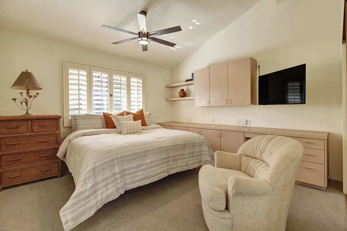 A bedroom with a bed, a chair, a dresser, a ceiling fan, a TV, and a window with blinds.