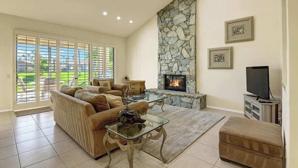 Una sala de estar con sofá, mesa de centro de vidrio, chimenea de piedra, televisor y ventanas grandes.