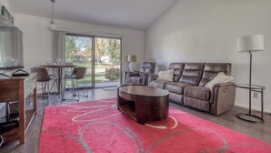 Una sala de estar moderna con un sofá de cuero marrón, una mesa de centro redonda y una alfombra roja. Hay un comedor con una mesa y sillas, y un ventanal que ofrece vistas al exterior.