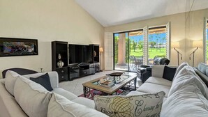 A modern living room with a sofa, coffee table, and TV stand. There is a sliding glass door leading to a patio with outdoor furniture. - Spacious-Dogs Ok-Great Views-VB887 (Palm Desert)