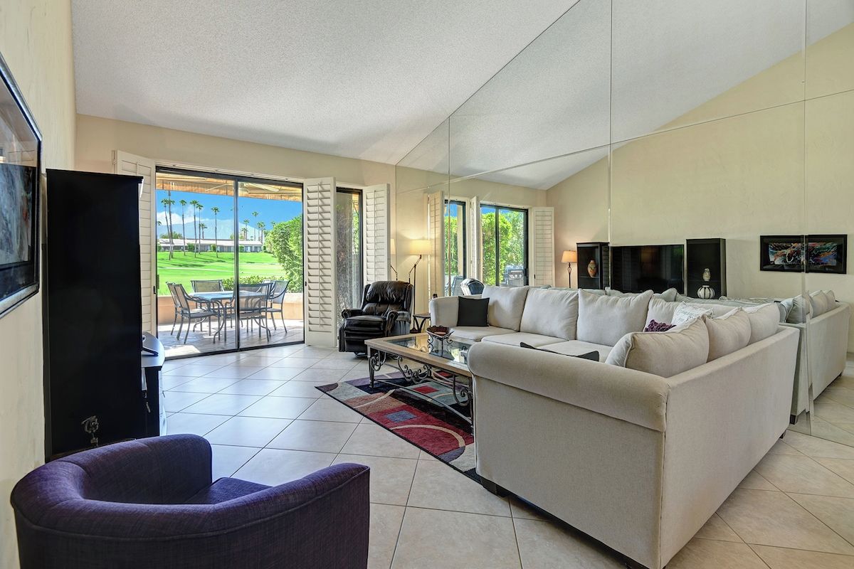 Step into your spacious living room with tile floors, plush seating, and French doors that open onto your private patio overlooking lush green lawns and towering palm trees.