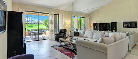 Step into your spacious living room with tile floors, plush seating, and French doors that open onto your private patio overlooking lush green lawns and towering palm trees.