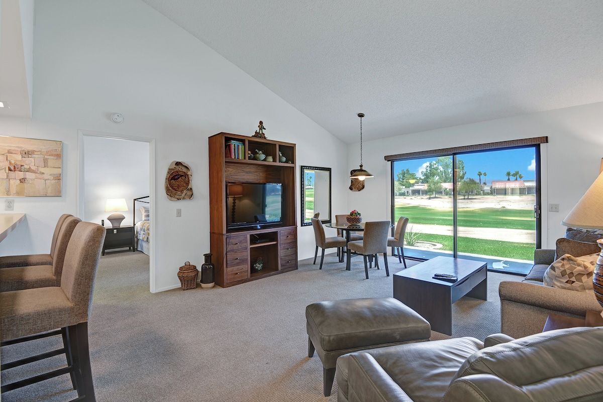Relax in your light-filled living space with comfortable seating, a cozy dining area, and sliding glass doors opening to views of palm trees and green lawns.