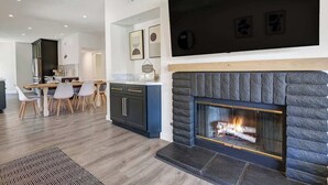 Een moderne woonkamer met een open haard, een eethoek met een houten tafel en stoelen, en een keuken op de achtergrond.