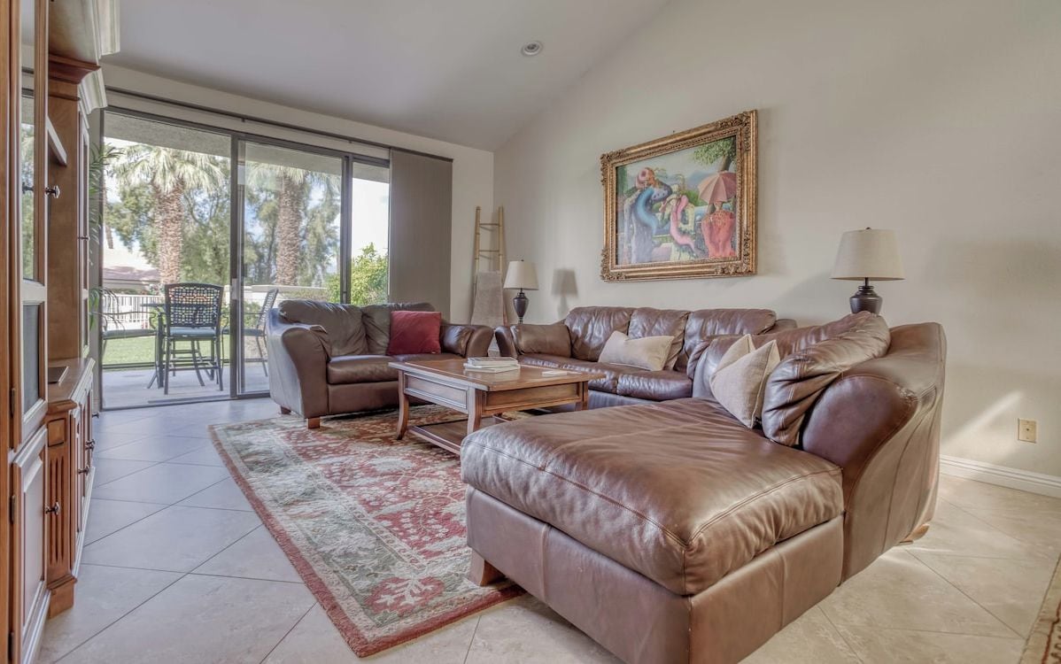 Sink into plush leather seating in your spacious living room, where sunlight streams through glass doors opening to a private patio surrounded by swaying palms and lush greenery.
