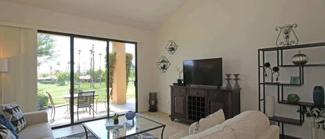 Sala de estar con sofá, mesa de centro y televisor, grandes puertas correderas de vidrio a un patio con mobiliario exterior.