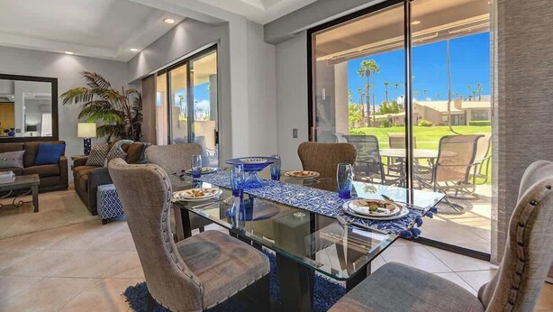 A dining area with a glass table and chairs, a blue patterned tablecloth, and a view of a green landscape through large sliding glass doors. - Walk to Pool and Spa-Patio-Fireplace-VB851 (Palm Desert)