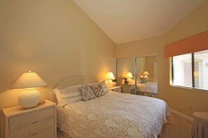 Ein Schlafzimmer mit Bett, Nachttischen, Lampen, einem Spiegel und einem Fenster mit Jalousien.