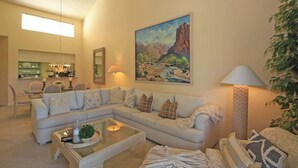 A living room with a sectional sofa, a glass coffee table, and a painting of a landscape on the wall. - Moutain and Fairway Views-Walk to Pool-V3986 (Palm Desert)