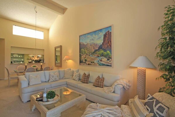A living room with a sectional sofa, a glass coffee table, and a painting of a landscape on the wall. - Moutain and Fairway Views-Walk to Pool-V3986 (Palm Desert)