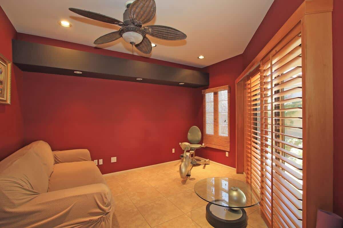 A room with a red wall, a ceiling fan, a glass table, a sofa, and a window with blinds.