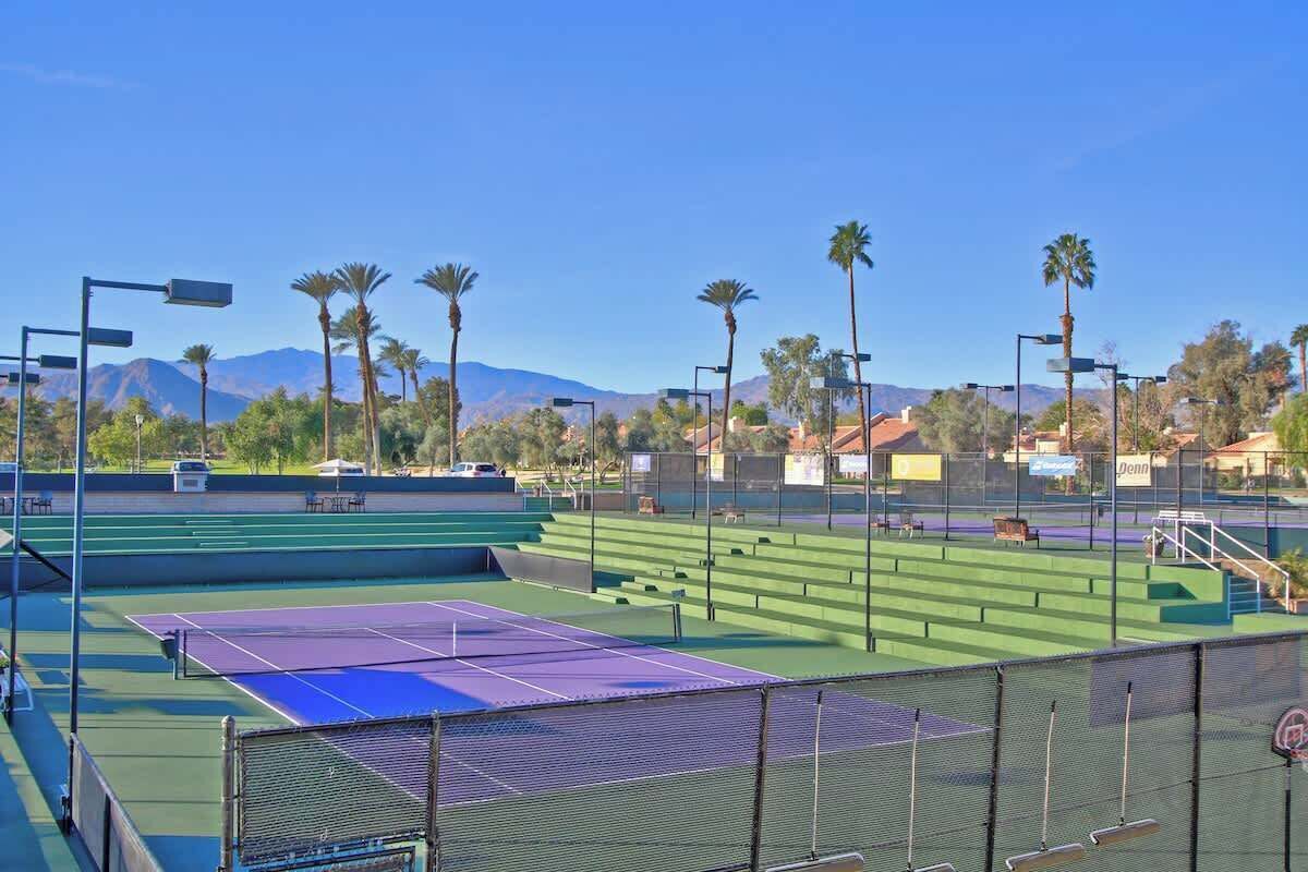 Un court de tennis avec un filet, entouré d’une clôture, et des palmiers en arrière-plan.