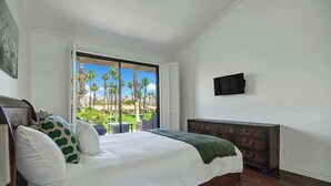 Un dormitorio con una cama grande, un aparador, un televisor montado en la pared y vistas a palmeras a través de un ventanal corredera.