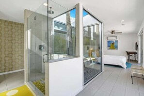 Et moderne hotelværelse med brusekabine i glas, seng, skrivebord og stol.
