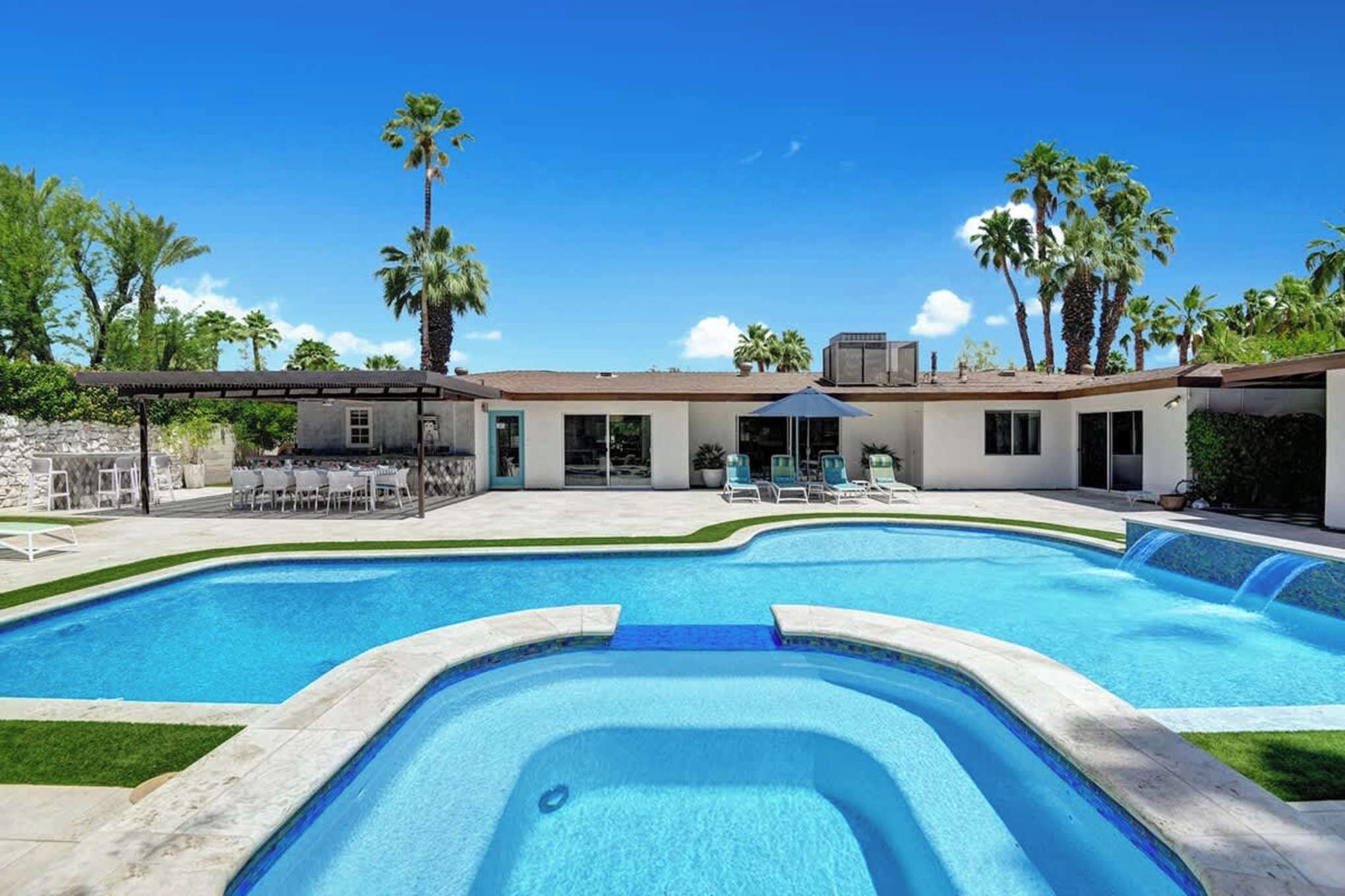 The property features a stunning desert oasis with an expansive pool area surrounded by lush palm trees under clear blue skies, offering an idyllic Palm Springs retreat setting.