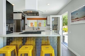 A modern kitchen with a large island, yellow stools, and a view of the outdoors. - Dog-Friendly Stunner|Pool|Fireplace|BBQ and more (Palm Springs)