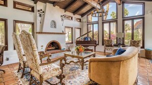 A spacious living room with a fireplace, a piano, and large windows offering views of the outdoors. - Pet-friendly tennis estate in The Mesa|360-views (Palm Springs)