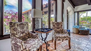 A sunlit room with two patterned armchairs, a small table with a board game, and large windows offering a view of the outdoors. - Pet-friendly tennis estate in The Mesa|360-views (Palm Springs)