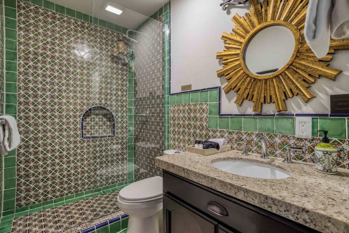 Een badkamer met groene mozaïektegels, een spiegel met gouden lijst, een wit toilet en een granieten aanrecht met een wastafel.
