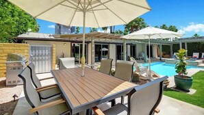 Une terrasse extérieure avec une table et des chaises en bois, un barbecue et des parasols pour faire de l’ombre.