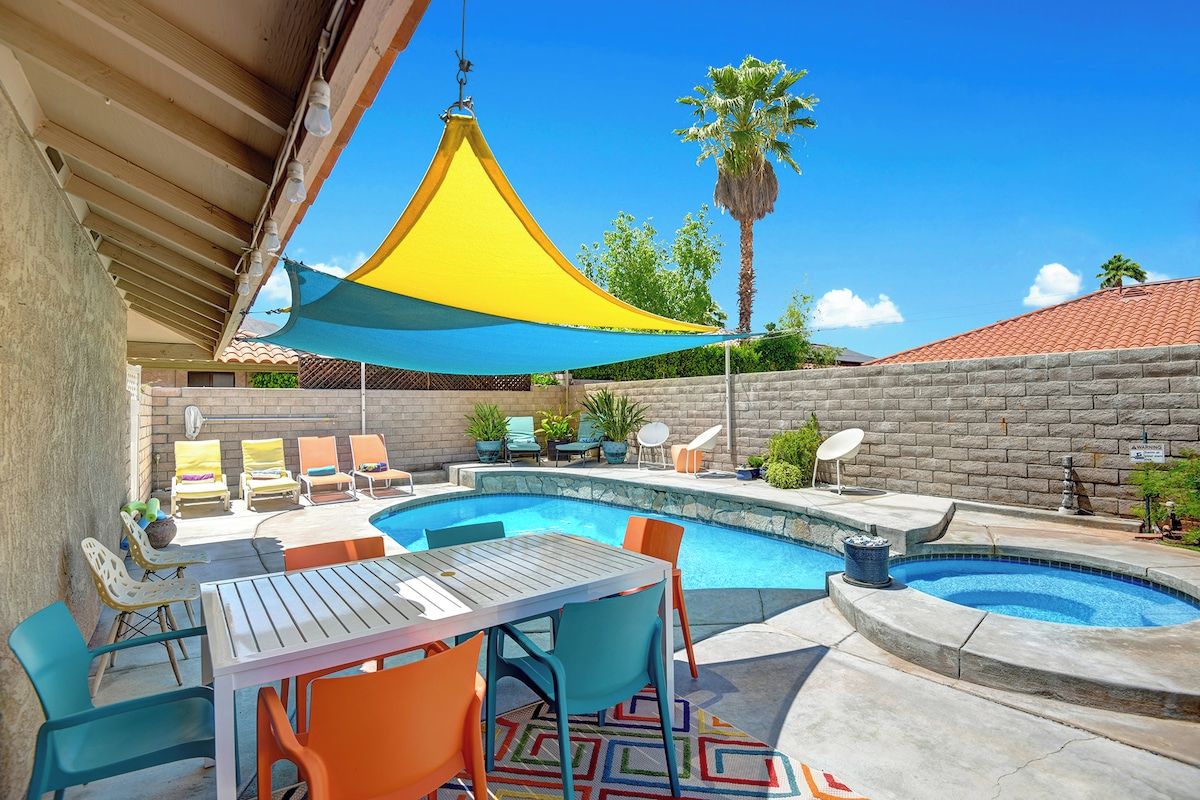 This property features a private backyard oasis with swimming pool, hot tub, covered patio dining, and colorful sun loungers perfect for enjoying the warm desert sunshine.