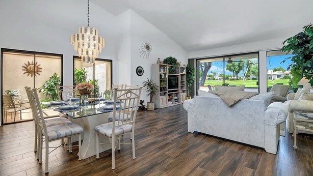Ein geräumiges Wohnzimmer mit einer großen weißen Couch, einem Essbereich mit einem Glastisch und Blick auf eine grüne Landschaft durch große Fenster.