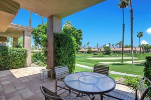 An outdoor patio with a round table and chairs, surrounded by greenery and palm trees, overlooking a well-maintained lawn and golf course. - Condo - The 7 Palm, Dogs OK, Fireplace , Pool (Palm Desert)