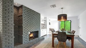 Ein modernes Wohnzimmer mit Kamin aus Backstein, einer Essecke mit Holztisch und -stühlen sowie einem großen Fenster mit Blick auf Grünflächen.