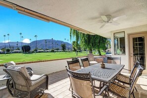 Une terrasse avec une table et des chaises, un ventilateur de plafond et une vue sur un champ verdoyant, avec des palmiers et des montagnes à l’horizon.