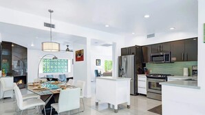 A modern kitchen with stainless steel appliances, a dining area with white chairs, and a large window with patterned curtains. - Teal and Tangerine Retreat - Movie Colony East - 5 mins to downtown, perfect 3 bedroom, 3 bath, Pool, Spa & Casita (Palm Springs)