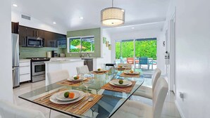A modern kitchen with stainless steel appliances, a glass dining table with placemats, and a view of a green garden through large windows. - Teal and Tangerine Retreat - Movie Colony East - 5 mins to downtown, perfect 3 bedroom, 3 bath, Pool, Spa & Casita (Palm Springs)
