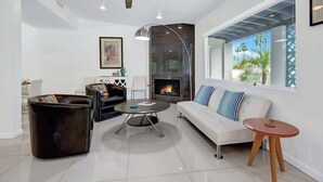A modern living room with a fireplace, a ceiling fan, and a large window with a view of palm trees. - Teal and Tangerine Retreat - Movie Colony East - 5 mins to downtown, perfect 3 bedroom, 3 bath, Pool, Spa & Casita (Palm Springs)