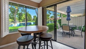 Un coin repas avec une table ronde et des tabourets, de grandes portes-fenêtres coulissantes donnant sur une terrasse aménagée avec du mobilier d’extérieur et offrant une vue sur une piscine.