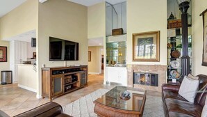 Un vaste salon comprenant un canapé en cuir, une table basse en verre, un téléviseur à écran plat, une cheminée et un coin cuisine avec des meubles blancs et un meuble vitrine en verre.