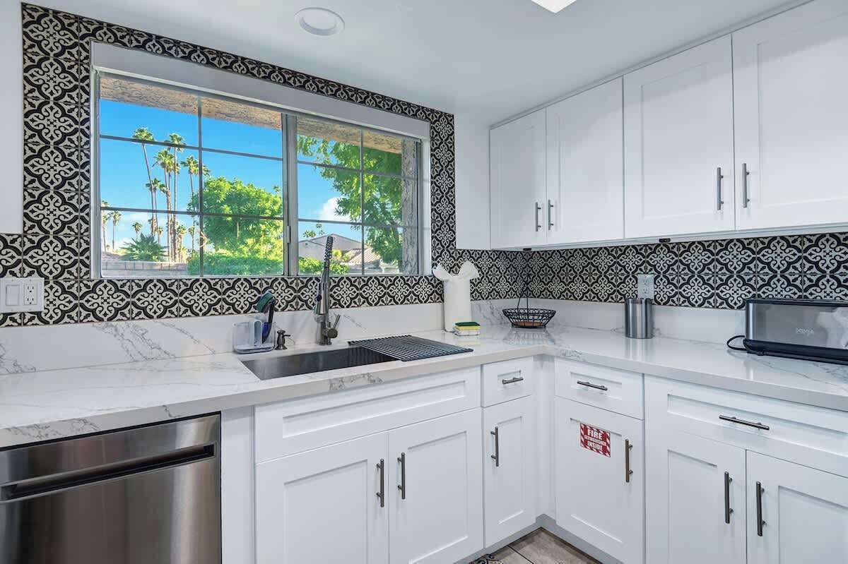 A modern kitchen with white cabinets, a stainless steel dishwasher, a sink with a black faucet, and a window with a view of palm trees.