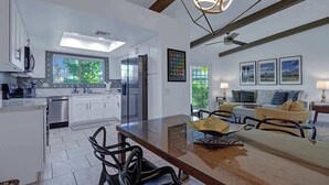 A modern kitchen with white cabinets, a dining area with a glass table, and a living room with a sofa and a TV. - Pet Friendly Bungalow in Mesquite Country Club (Palm Springs)