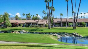A golf course with a pond, palm trees, and a building with a tiled roof. - Pet Friendly|2 Suites+ guest room|Large Patio|Fairway Views (Palm Desert)