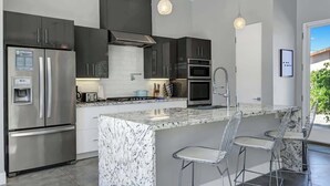 A modern kitchen with dark cabinets, stainless steel appliances, a marble island, and bar stools. - Pet-friendly Desert Park home with heated pool|Spa (Palm Springs)