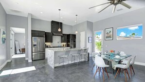 A modern kitchen with a central island, stainless steel appliances, and a dining area with white chairs and a glass table. - Pet-friendly Desert Park home with heated pool|Spa (Palm Springs)
