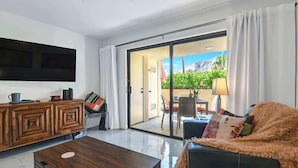 Een moderne woonkamer met een flatscreen-tv, een houten kast, een bank en een schuifpui die leidt naar een buitenruimte met een tafel en stoelen.