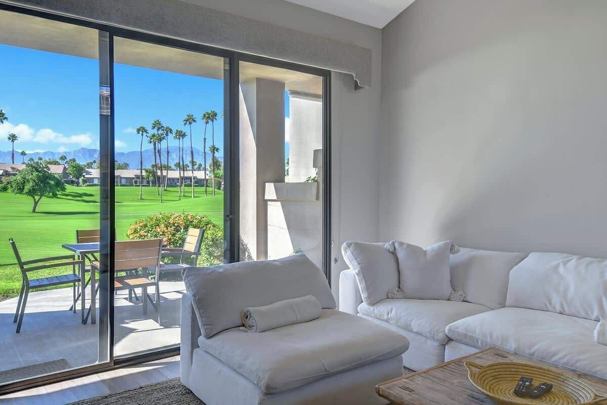 A modern living room with a white sofa, a glass door leading to a balcony with a view of a golf course and palm trees.