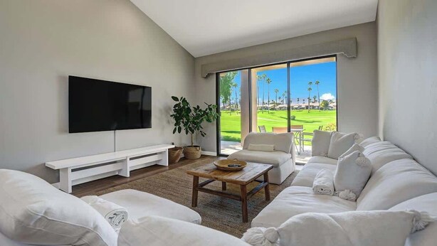 A modern living room with a large flat-screen TV, white sofas, a wooden coffee table, and a large window offering a view of a golf course. - Fairway View Escape in Palm Desert (Palm Desert)