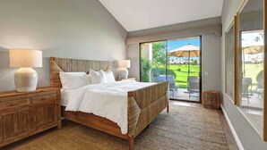 A bedroom with a large bed, wooden nightstands, two bedside lamps, and a view of a garden through a sliding glass door. - Fairway View Escape in Palm Desert (Palm Desert)
