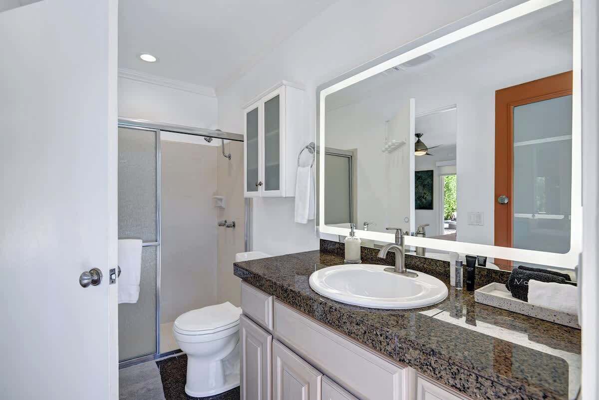 A modern bathroom with a glass shower enclosure, a white toilet, a granite countertop sink, and a large mirror above the sink.