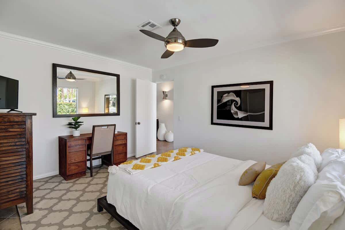 A bedroom with a bed, a desk, a chair, a ceiling fan, a mirror, and a framed picture on the wall.